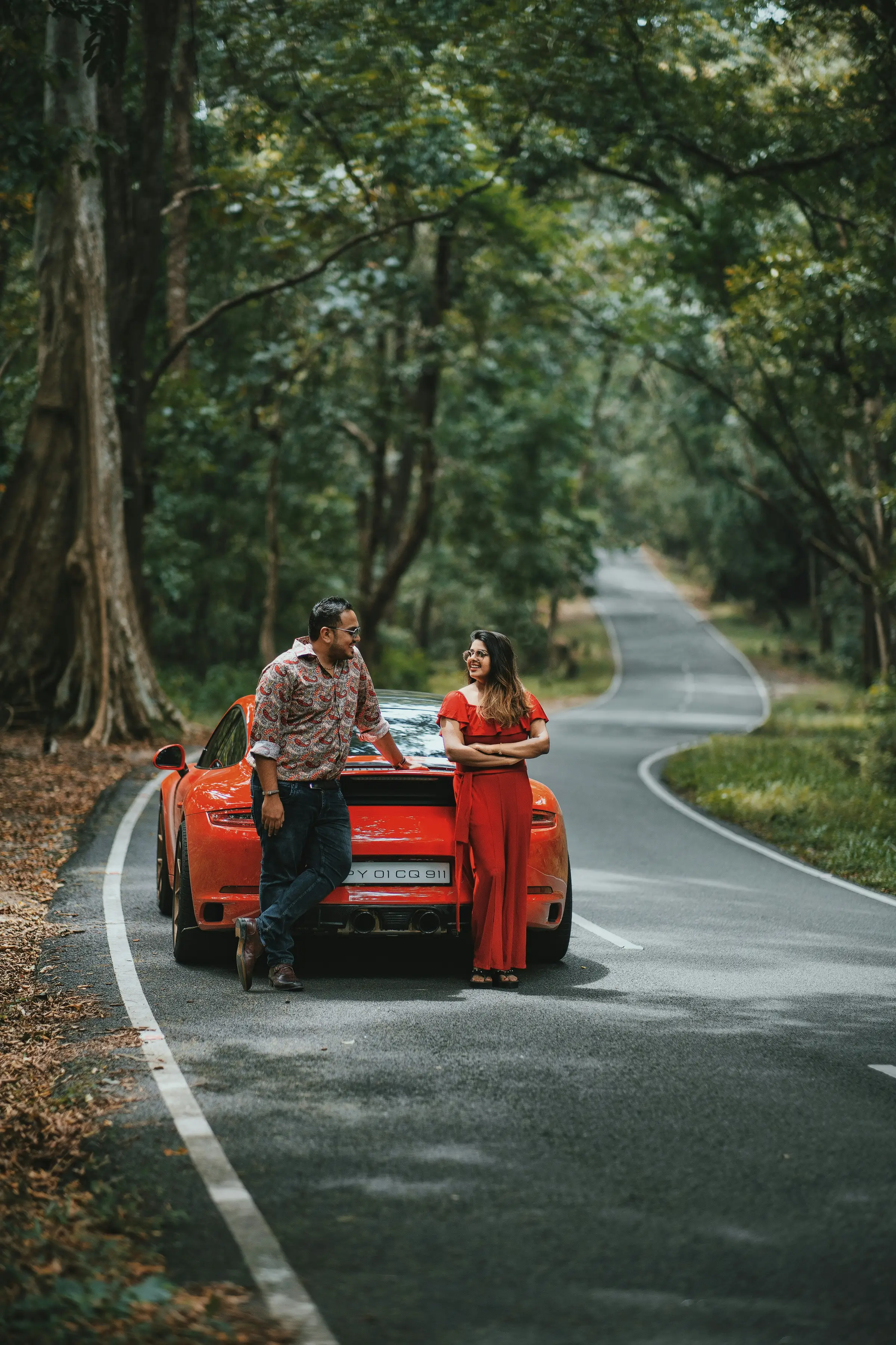 A couple leaning against their Porsche 911 on a tree-lined road, sharing a private luxury driving tour
